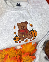 A light heather gray sweatshirt featuring an embroidered autumn scene with a brown teddy bear wearing a red scarf, sitting between two pumpkins and surrounded by orange and yellow leaves. The sweatshirt is styled with real orange and yellow autumn leaves.