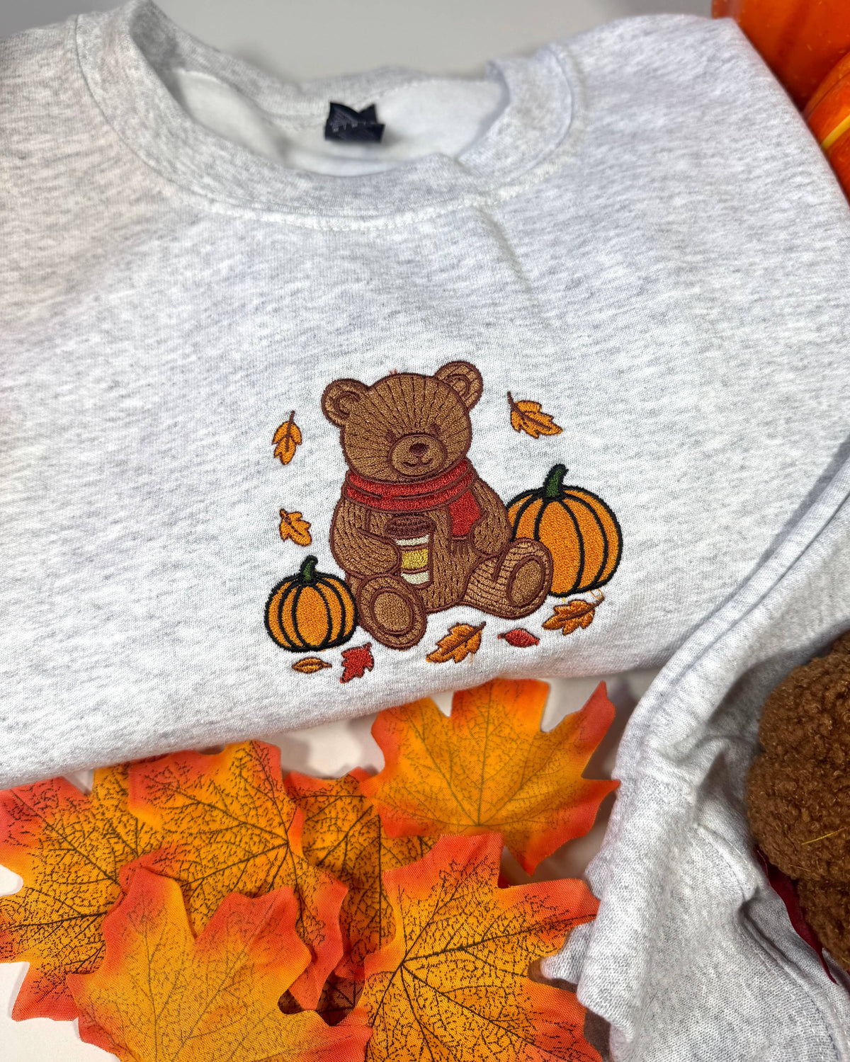 A light heather gray crewneck sweatshirt featuring a central autumn-themed embroidery of a brown teddy bear wearing a red scarf, sitting between two orange pumpkins with scattered fall leaves. The sweatshirt is styled with orange and yellow autumn leaves and a pumpkin in the background.