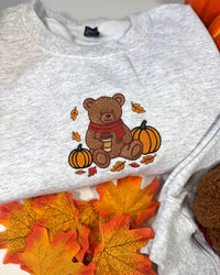 A light heather gray crewneck sweatshirt featuring a central autumn-themed embroidery of a brown teddy bear wearing a red scarf, sitting between two orange pumpkins with scattered fall leaves. The sweatshirt is styled with orange and yellow autumn leaves and a pumpkin in the background.