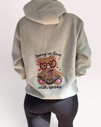 A person shown from the back wearing a light beige hoodie with a large graphic of a teddy bear wearing glasses and holding an open book, surrounded by small red hearts and the text 'beary in love with books'. The person is also wearing dark gray leggings against a plain light background.