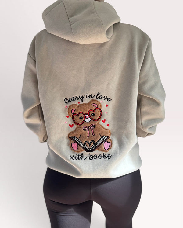 A person shown from the back wearing a light beige hoodie with a large graphic of a teddy bear wearing glasses and holding an open book, surrounded by small red hearts and the text 'beary in love with books'. The person is also wearing dark gray leggings against a plain light background.