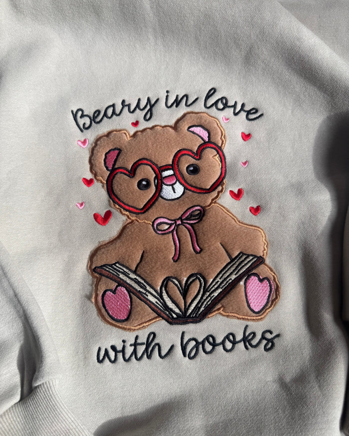 A close-up of a light gray sweatshirt featuring a detailed embroidered design of a brown teddy bear wearing heart-shaped glasses and holding an open book. The text 'Beary in love with books' is embroidered around the bear, accented with small red hearts.
