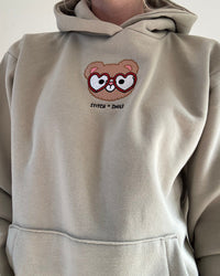 A person wearing a light beige hoodie with a small embroidered cartoon bear face wearing heart-shaped glasses on the chest, with the text 'STITCH + SHOE!' below it.