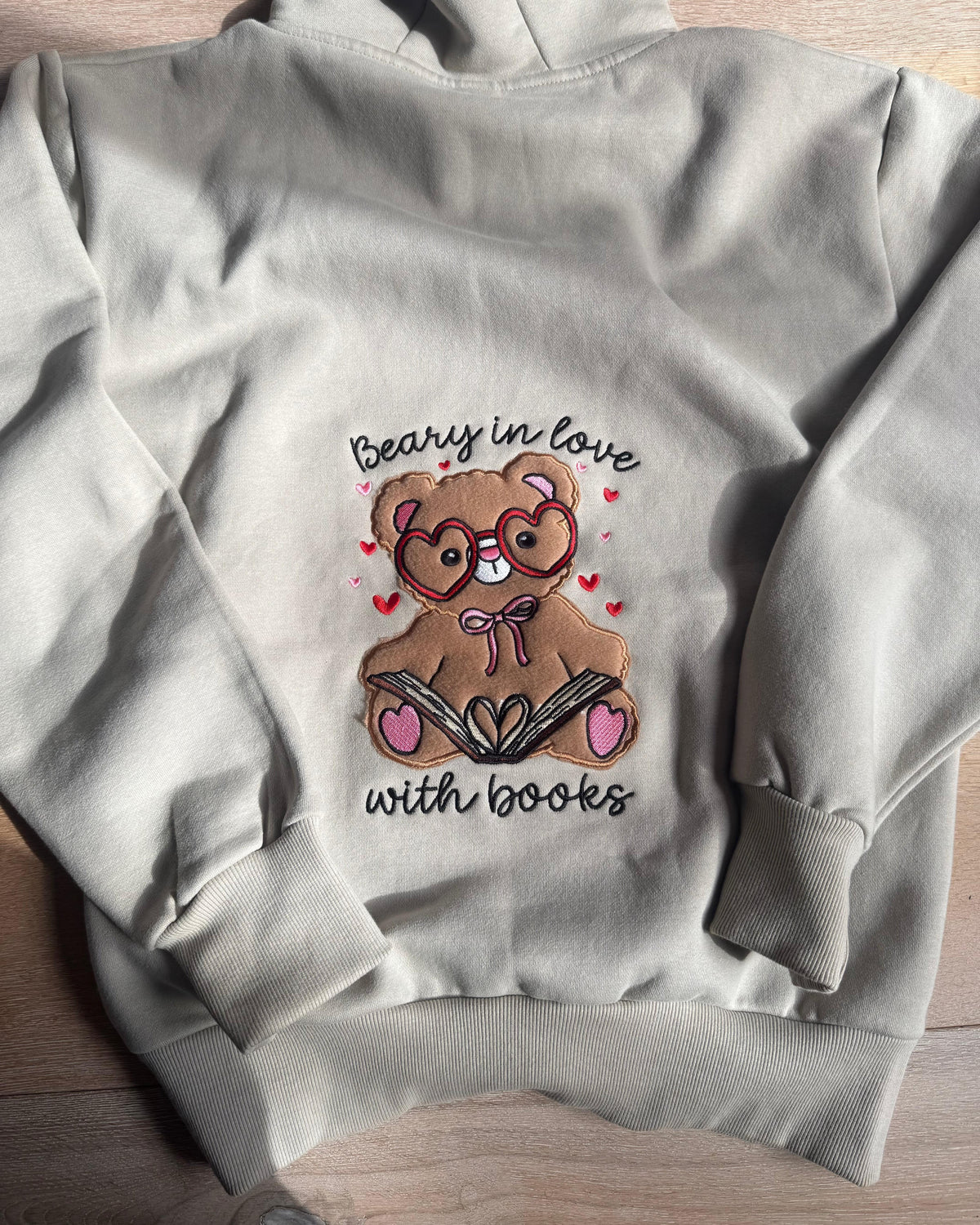 A light gray hoodie laid flat on a light wood surface, featuring a central graphic of a brown teddy bear wearing heart-shaped glasses and reading a book, surrounded by small pink hearts, with the text 'Beary in love with books' in black script.