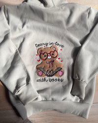 A light gray hoodie laid flat on a light wood surface, featuring a central graphic of a brown teddy bear wearing heart-shaped glasses and reading a book, surrounded by small pink hearts, with the text 'Beary in love with books' in black script.