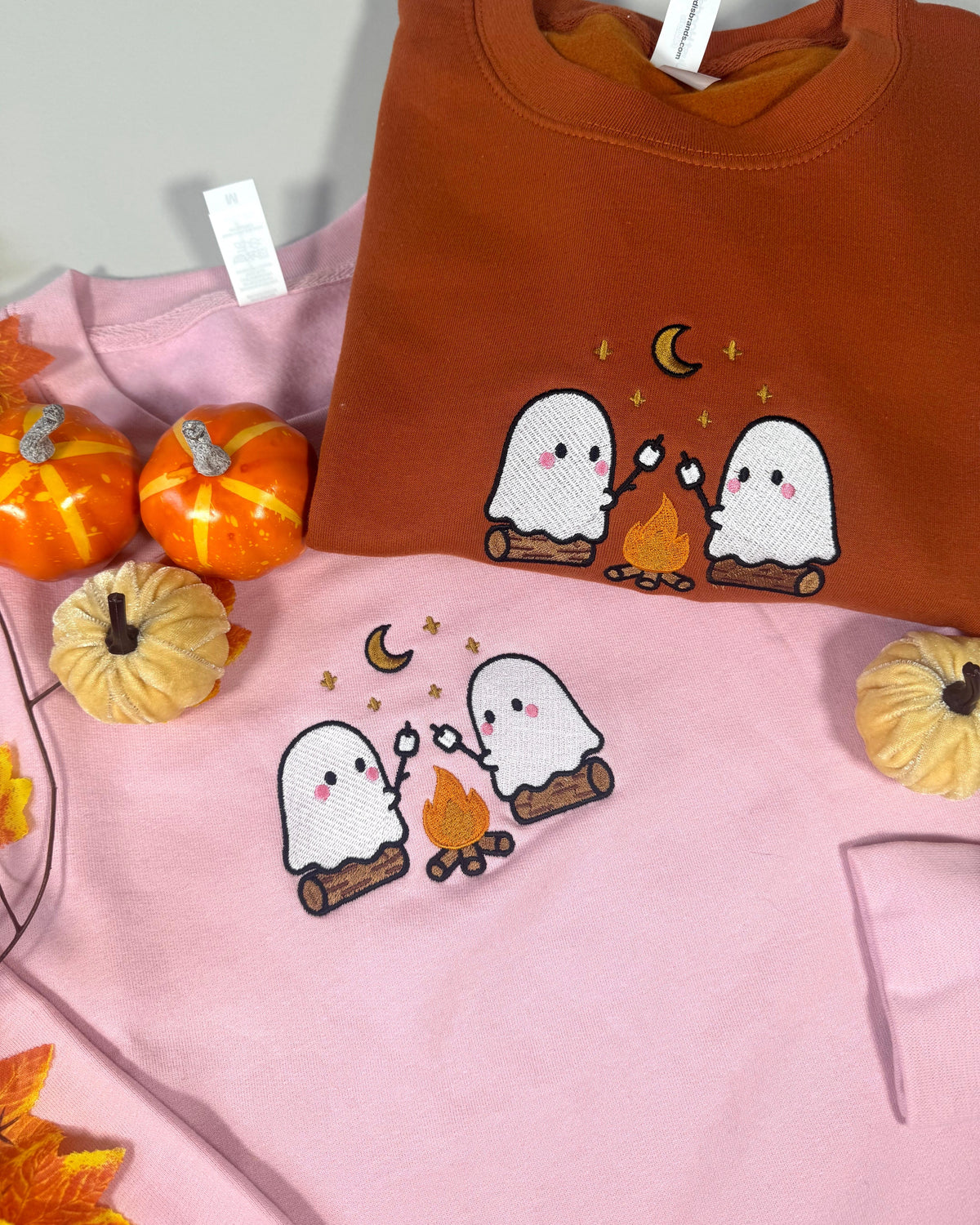 Two crewneck sweatshirts displayed together with decorative pumpkins and autumn leaves. The top sweatshirt is rust orange, and the bottom sweatshirt is light pink, both featuring a cute embroidered design of two white ghosts roasting over a small campfire under a crescent moon.