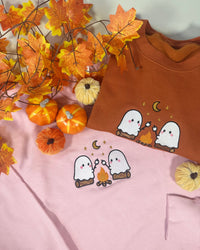 A flat-lay of two children's long-sleeve shirts, one in rust brown and one in light pink, both featuring a cute graphic of two white ghosts hugging around a small campfire with stars and a crescent moon. The shirts are styled with autumn-themed props including artificial maple leaves and small pumpkins on a light gray background.