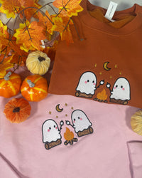 Two orange crewneck sweatshirts displayed on a pink surface with autumn props, including orange pumpkins and artificial maple leaves. Each sweatshirt features a cute embroidered design of two white ghosts sitting by a campfire under a crescent moon and stars.