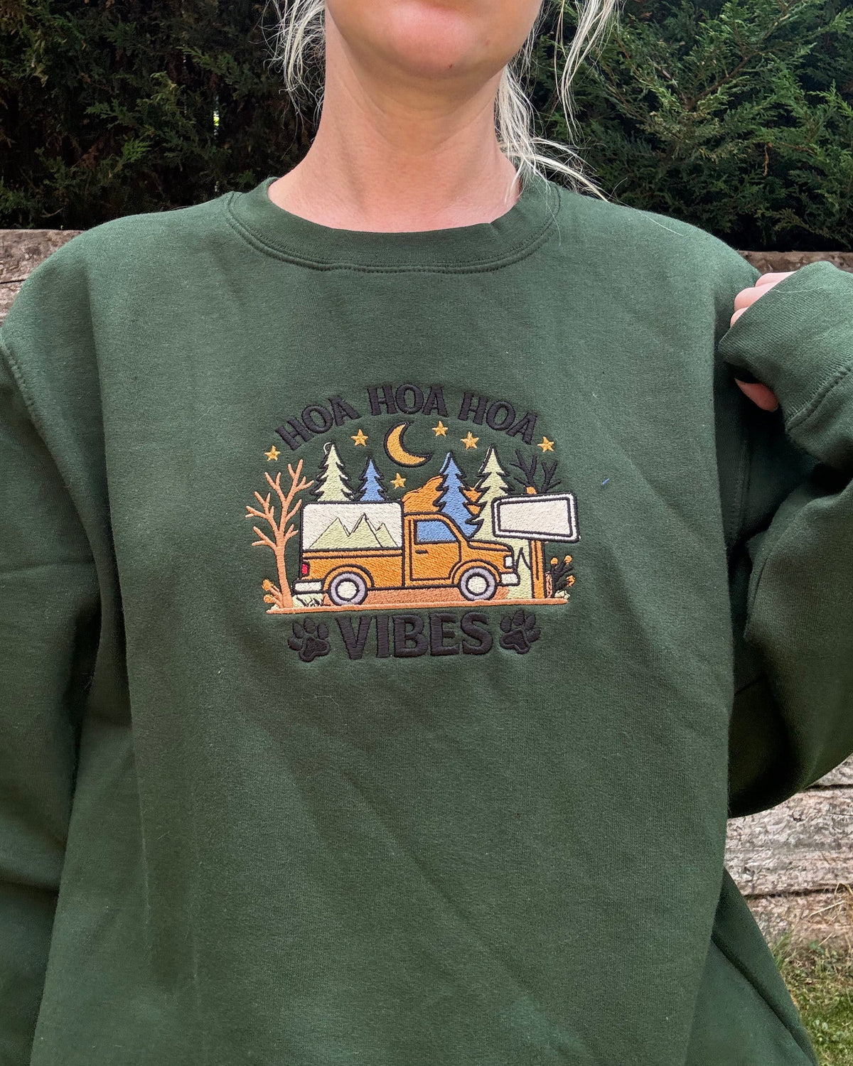 A person wearing an olive green crewneck sweatshirt with a colorful graphic on the chest depicting a camping scene and the text 'HOW HOY A DAY VIBES'.
