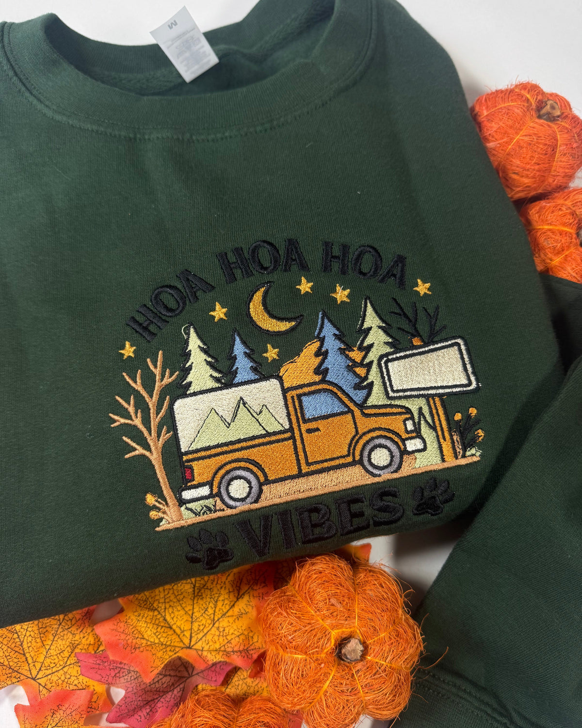 A dark green crewneck sweatshirt featuring a colorful embroidered graphic of an orange camp truck under a crescent moon and stars, with the text 'HOA HOA HOA VIBES' and paw prints. The sweatshirt is styled with autumn-themed yarn pumpkins and leaves on a white background.