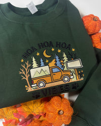 A dark green crewneck sweatshirt featuring a colorful embroidered graphic of an orange camp truck under a crescent moon and stars, with the text 'HOA HOA HOA VIBES' and paw prints. The sweatshirt is styled with autumn-themed yarn pumpkins and leaves on a white background.