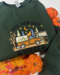 A dark forest green crewneck sweatshirt featuring a colorful embroidered graphic of an orange pickup truck in a pine forest at night under a crescent moon and stars, with the text 'HOA HOA HOA VIBES' embroidered around it. The sweatshirt is styled with autumn-themed props including orange pumpkins and fall leaves.