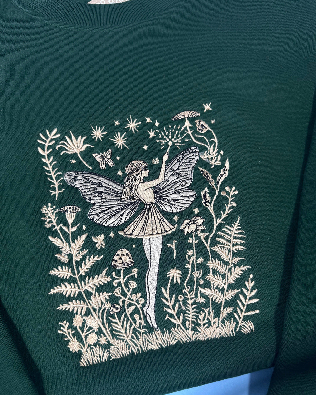  This premium embroidered sweatshirt features a beautifully detailed fairy illustration surrounded by delicate wildflowers, mushrooms, and stars