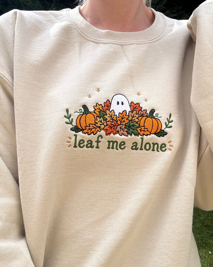 A person wearing a cream-colored crewneck sweatshirt with a fall-themed graphic on the chest featuring pumpkins, autumn leaves, a small ghost, and the text 'leaf me alone'.