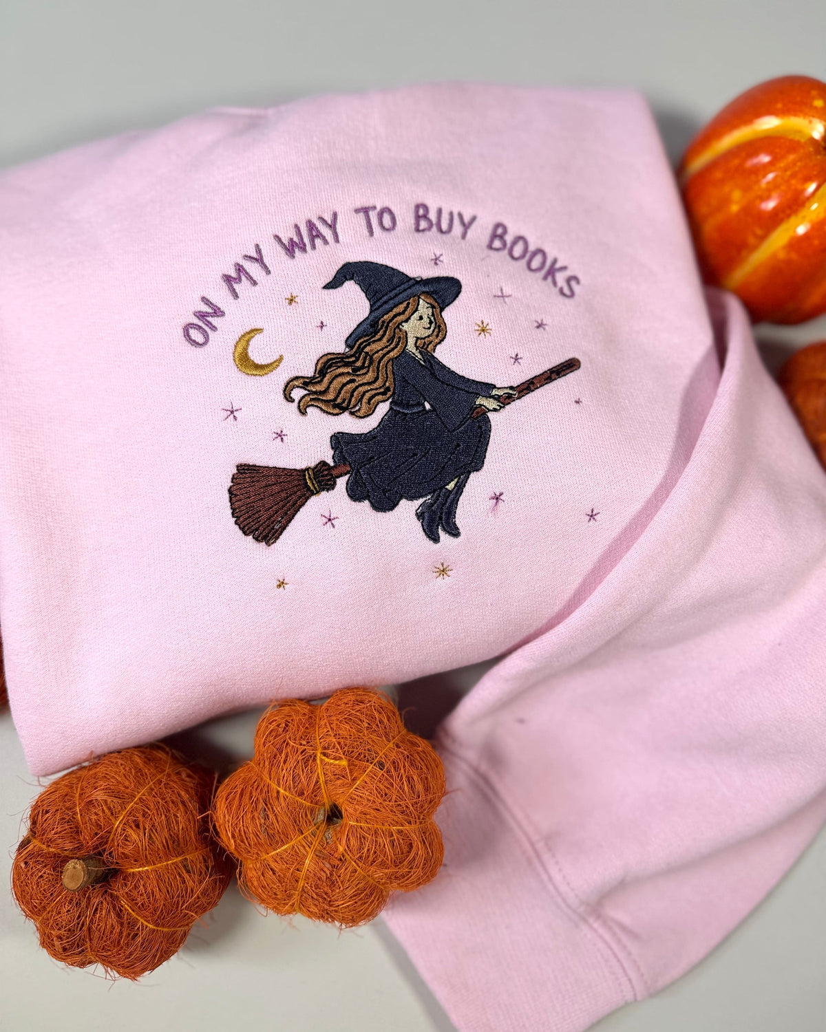 A folded light pink sweatshirt with an embroidered design of a witch riding a broomstick and the text 'ON MY WAY TO BUY BOOKS' in purple. The sweatshirt is styled with small orange decorative pumpkins on a light gray background.