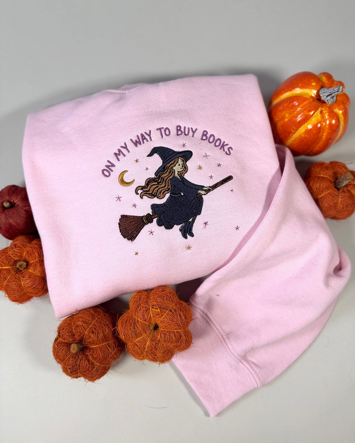 A folded light pink sweatshirt with a Halloween-themed embroidered design featuring a witch on a broomstick and the text 'ON MY WAY TO BUY BOOKS'. The sweatshirt is styled with small decorative pumpkins on a neutral gray background.
