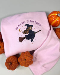 A folded light pink sweatshirt with an embroidered design of a witch riding a broomstick and the text 'ON MY WAY TO BUY BOOKS', styled with small orange pumpkins on a light gray background.